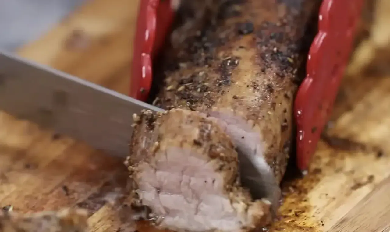 Un jugoso solomillo de cerdo al horno, recién cortado en una tabla de madera. Las pinzas rojas sujetan la carne mientras un cuchillo la rebana.
