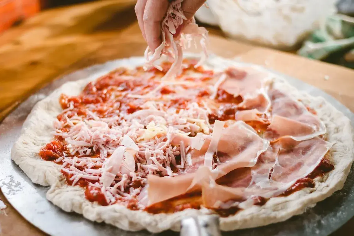 Manos esparciendo queso rallado sobre una pizza casera con jamón y salsa de tomate.
