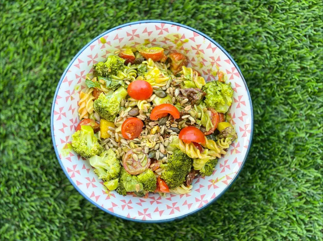 Deliciosa ensalada de pasta con brócoli, tomates cherry y semillas, servida en un cuenco con estampado geométrico sobre césped verde.