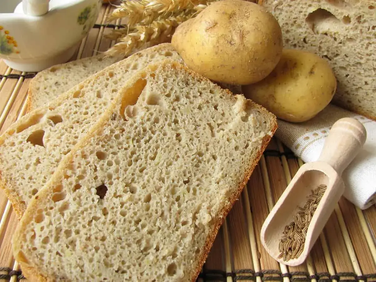 Rebanadas de pan de patata rústico, con patatas enteras, espigas de trigo y comino en una cuchara de madera.