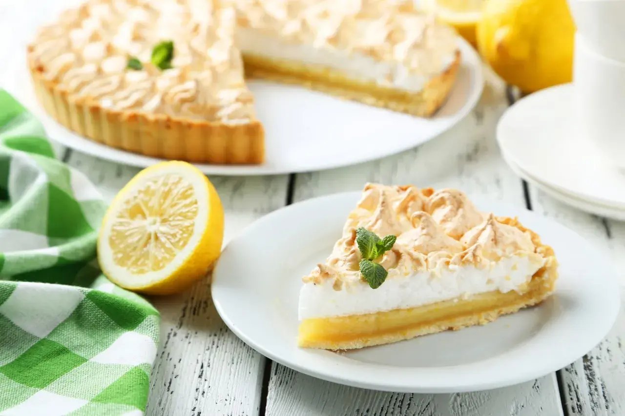 Delicioso pastel de limón con merengue dorado, decorado con menta fresca. Un trozo servido en plato blanco, junto a limones y una taza de té.
