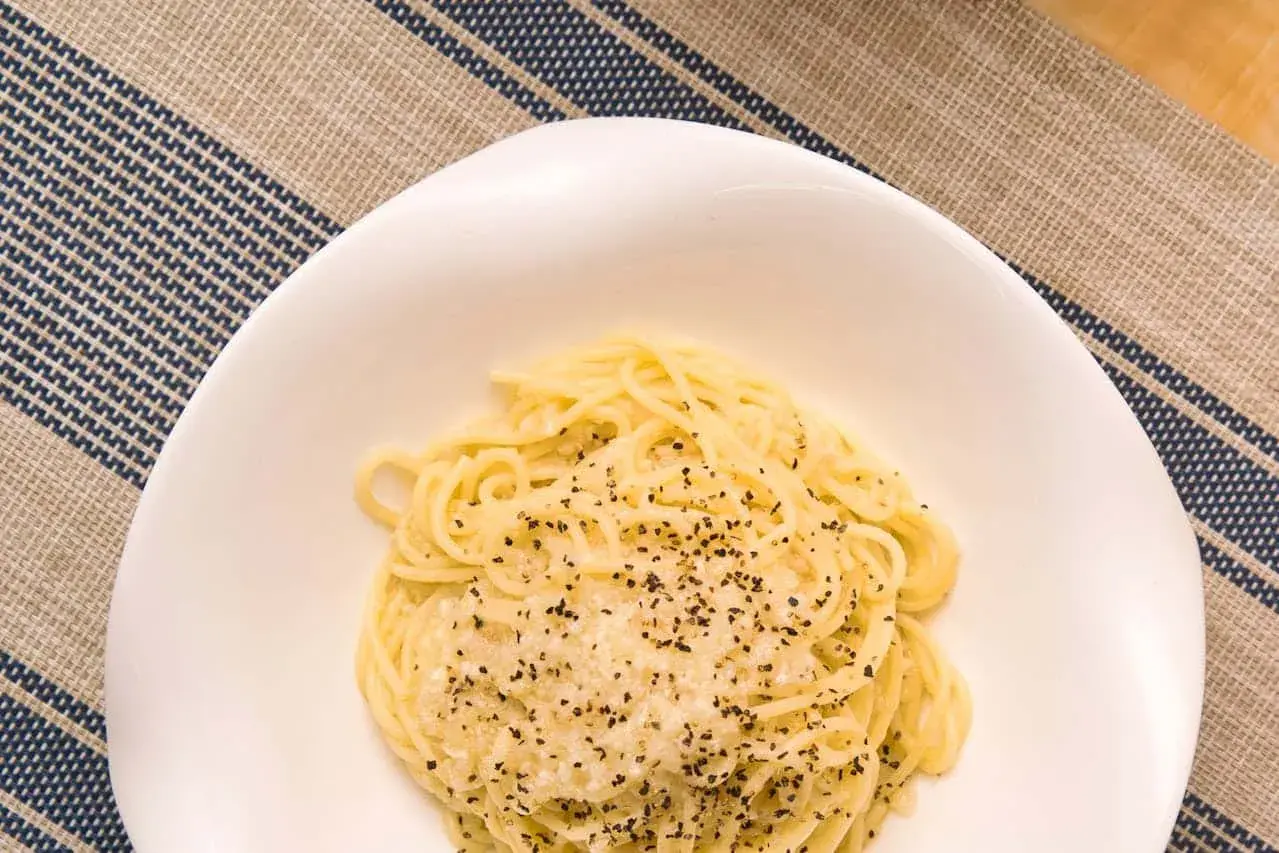 Un plato de cacio e pepe, pasta cremosa con queso y pimienta negra, servido en un cuenco blanco sobre un mantel a rayas.