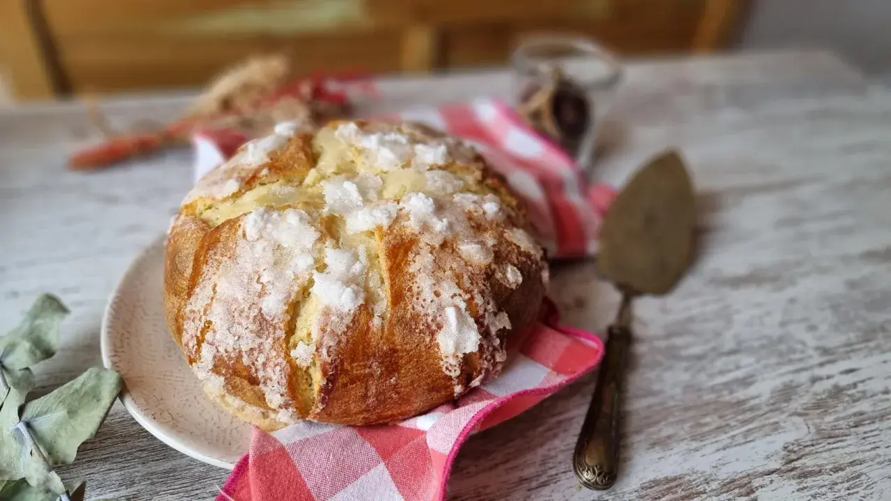 Un pan quemado dorado y crujiente, espolvoreado con azúcar, descansa sobre un plato. Una servilleta a cuadros rojos y blancos y un utensilio para pasteles completan la escena.