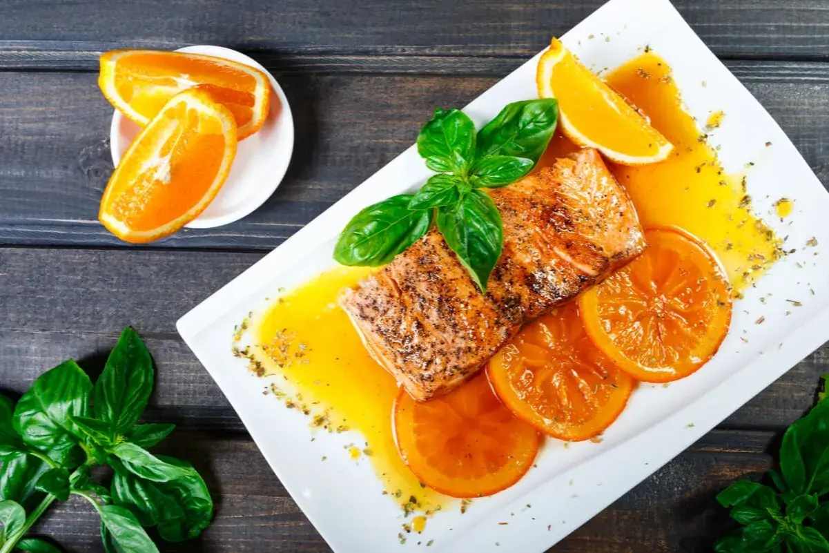 Delicioso salmón a la naranja, servido con rodajas de naranja y hojas de albahaca fresca.