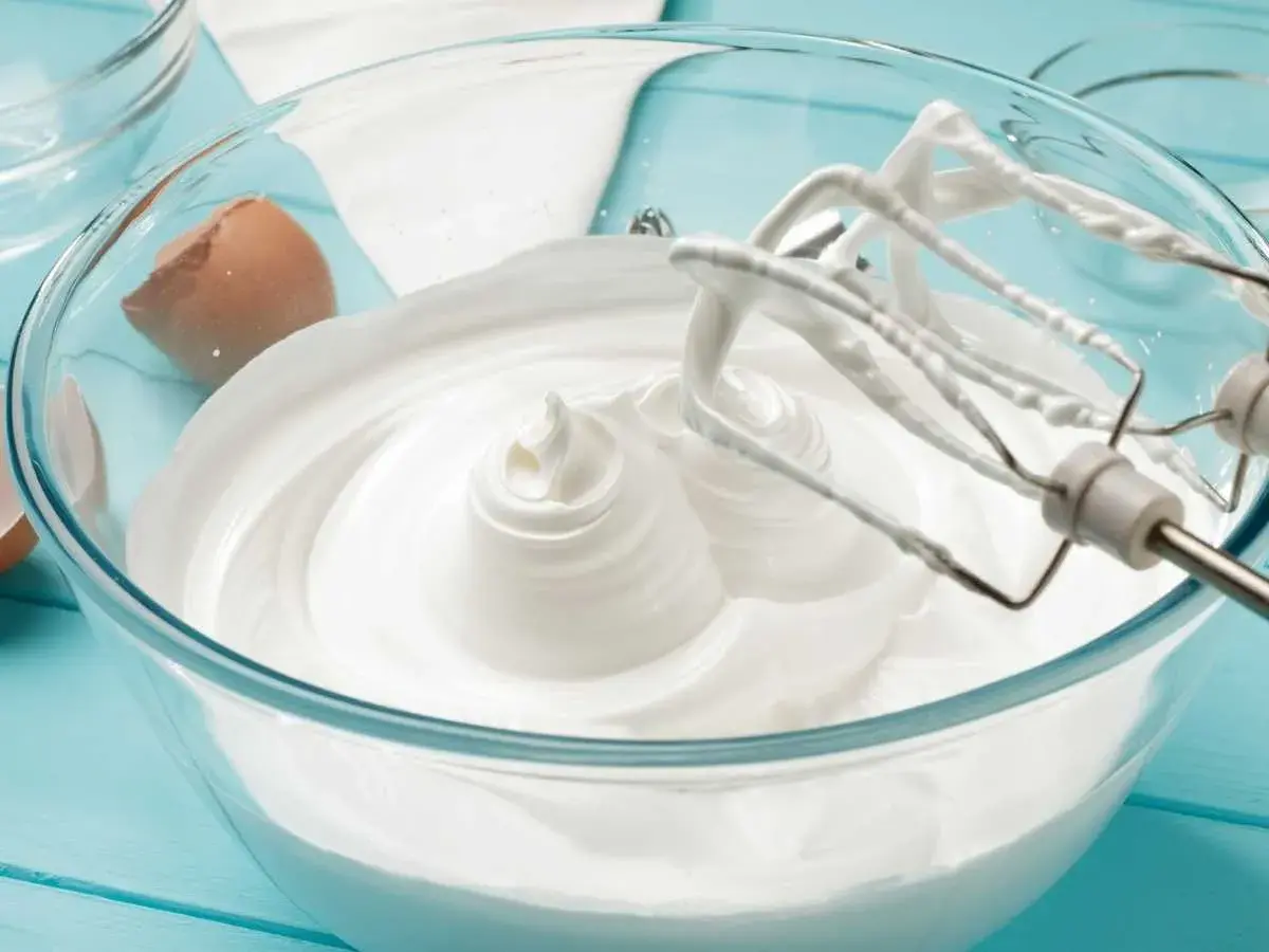 Batidora mezclando merengue italiano en un bol de cristal. Cáscara de huevo al lado.
