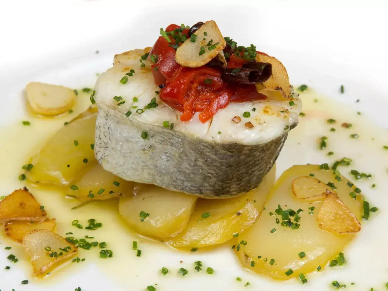 Deliciosa merluza a la plancha sobre patatas, con pimientos asados, ajo crujiente y perejil picado.