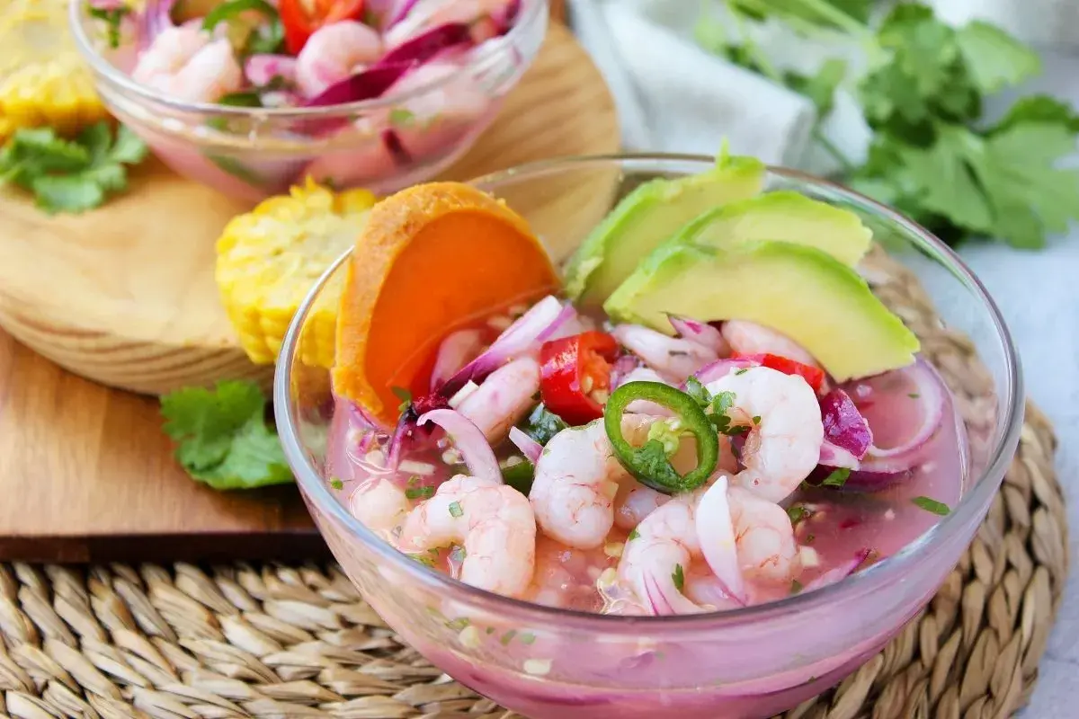 Delicioso ceviche de gambas con aguacate, cebolla morada, chile jalapeño y tomate. Servido en un cuenco de cristal, acompañado de maíz y cilantro fresco.