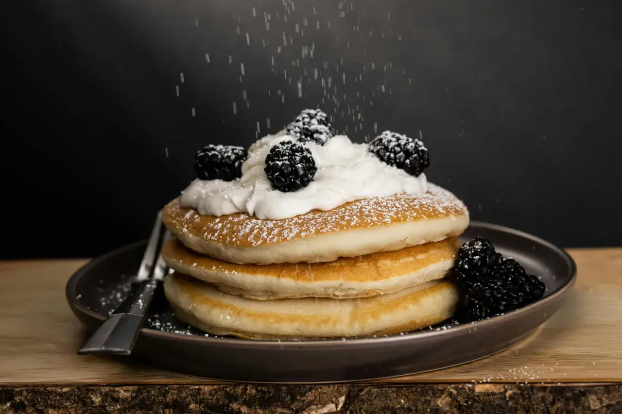 Una pila de tortitas americanas cubiertas con crema batida y moras, espolvoreadas con azúcar glas.