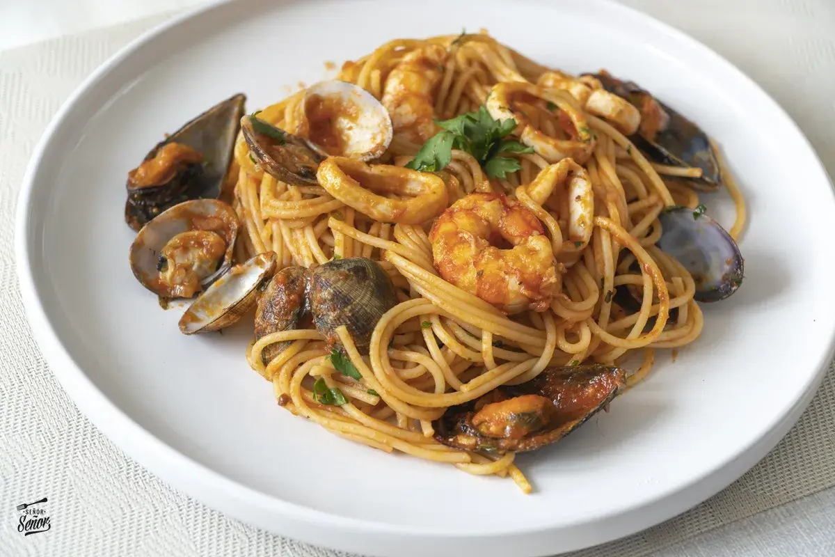 Espaguetis a la marinera con gambas, mejillones, almejas y calamares, servidos en un plato blanco.