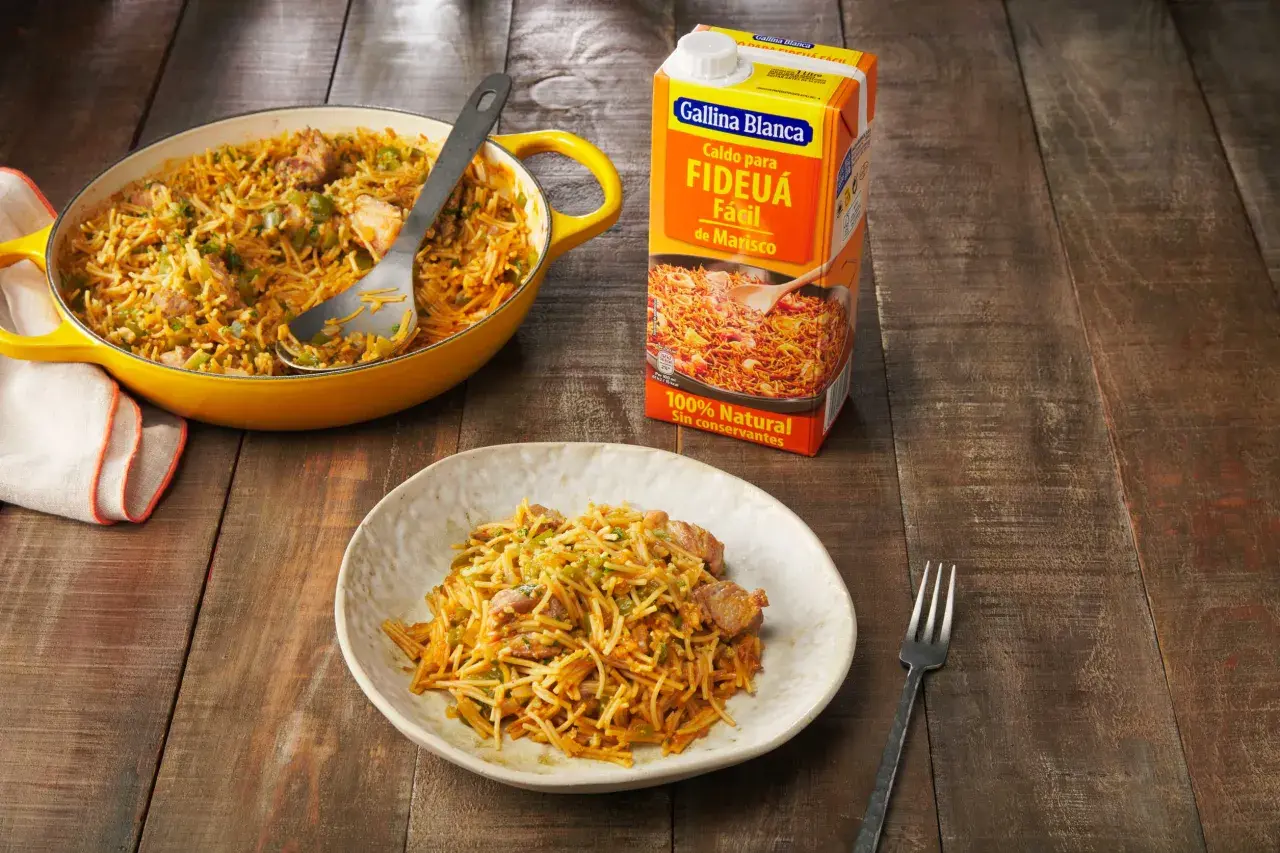 Fideos a la cazuela servidos en un plato y una paellera amarilla, junto a un caldo para fideuá.