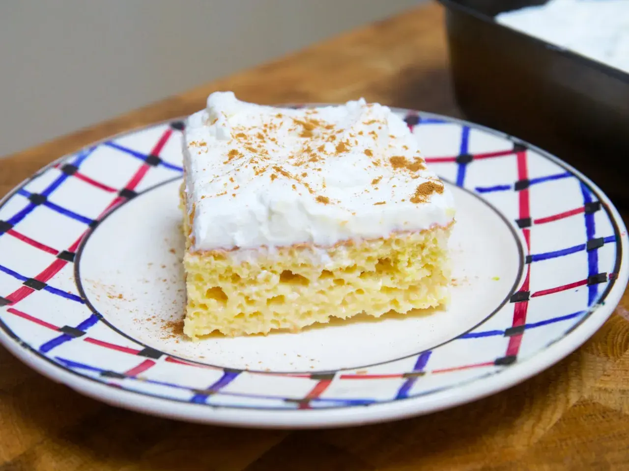 Un trozo de tarta tres leches, esponjoso y cubierto de crema batida con canela, servido en un plato decorado.