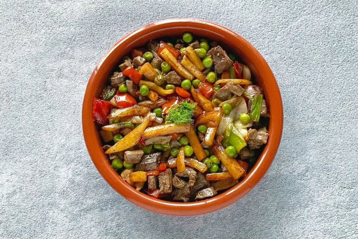 Un sabroso frito mallorquín con trozos de carne, patatas fritas, guisantes y pimientos rojos en una cazuela de barro.