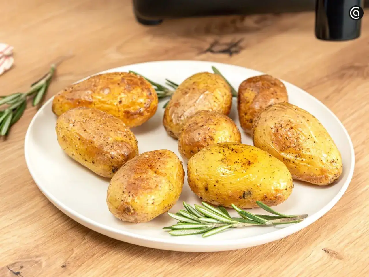 Patatas asadas en freidora de aire, doradas y crujientes, servidas con romero fresco en un plato blanco sobre una mesa de madera.