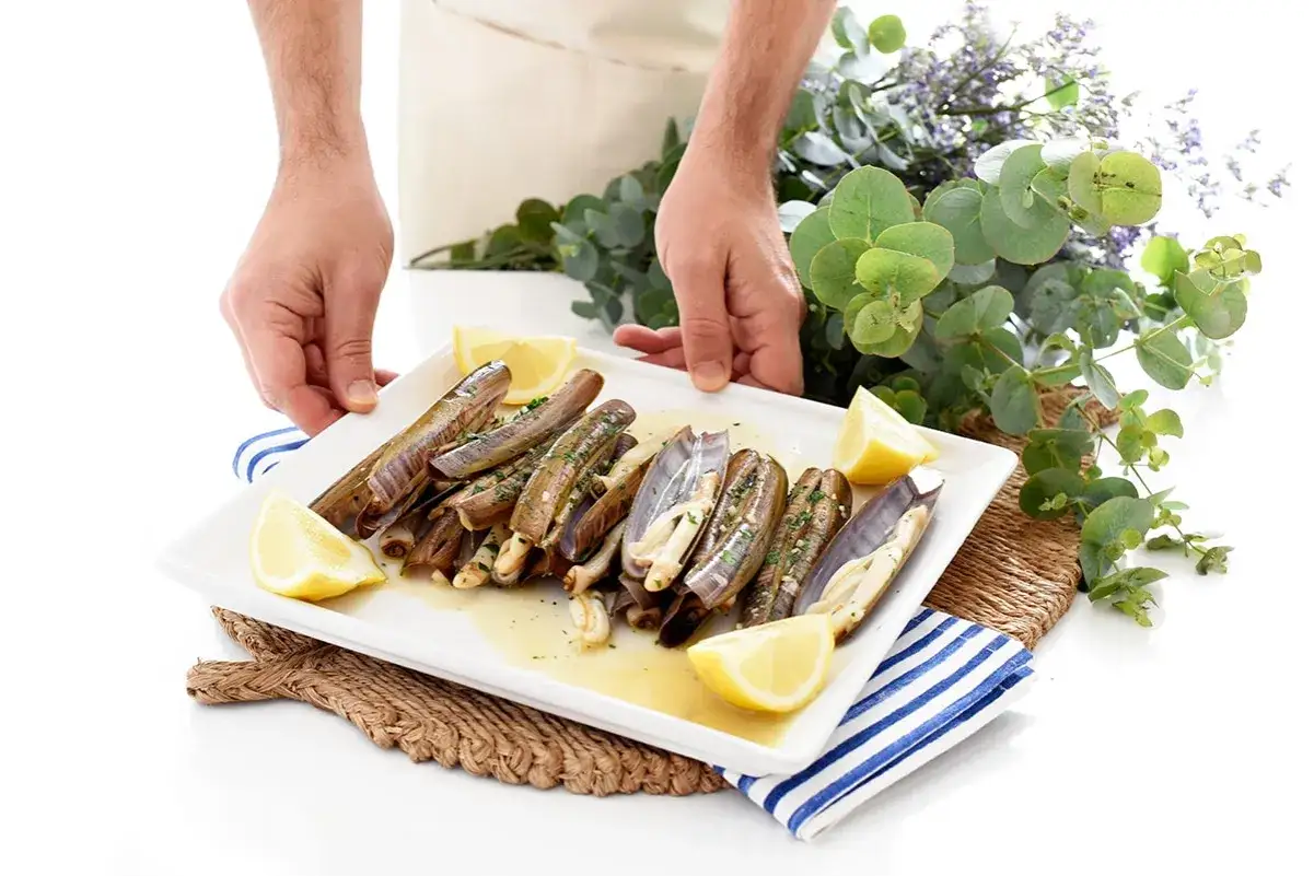 Manos sirviendo un plato de navajas a la plancha, adornado con gajos de limón y perejil picado.