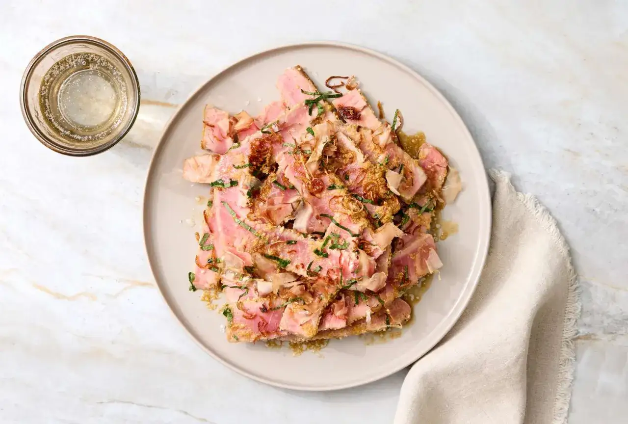 Tataki de at&uacute;n fresco, cortado en l&aacute;minas finas y adornado con hierbas y crujientes. Servido con una copa de bebida espumosa.