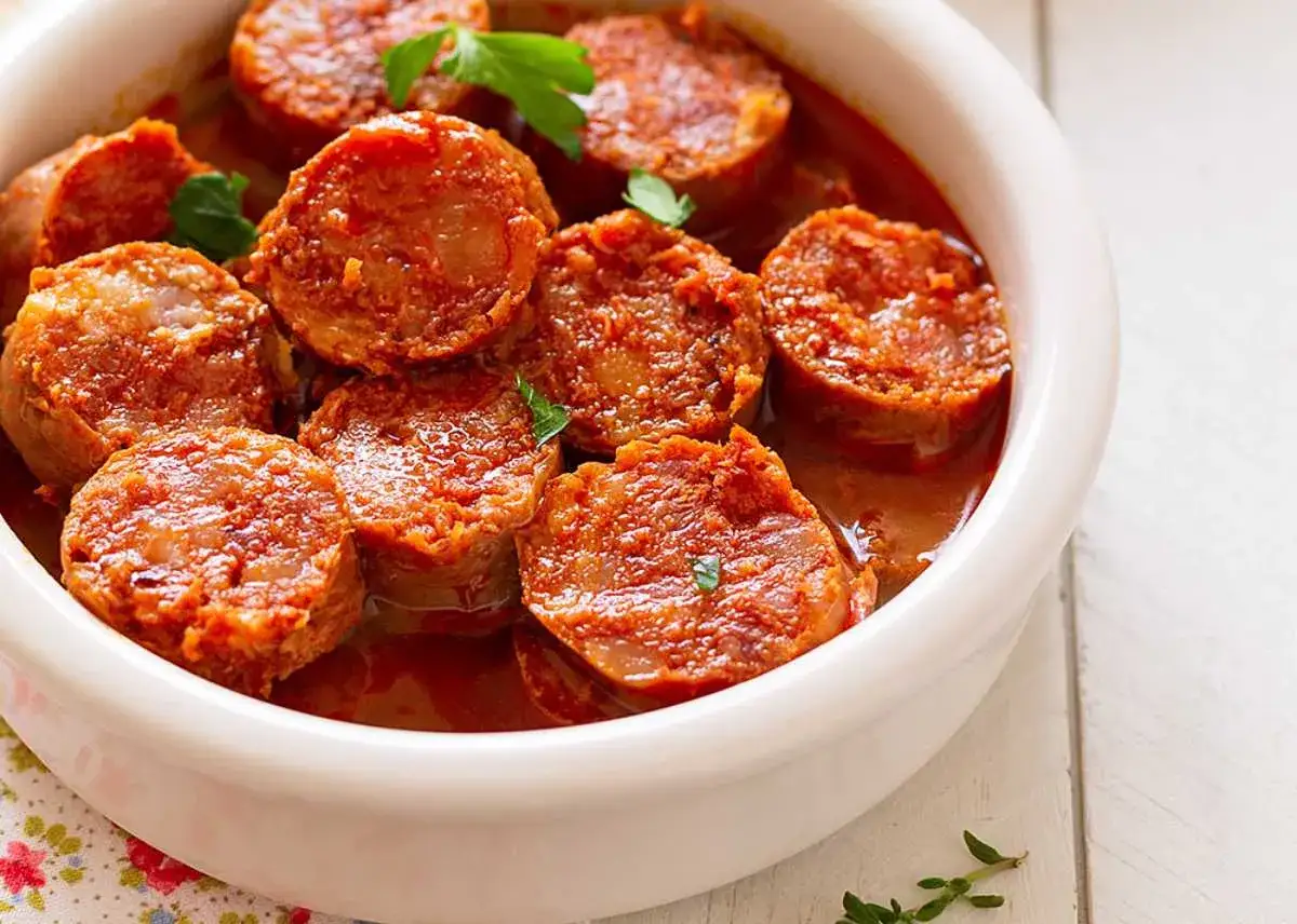 Chorizo a la sidra, jugosas rodajas de chorizo cocinadas en una salsa rojiza, servidas en un cuenco blanco.