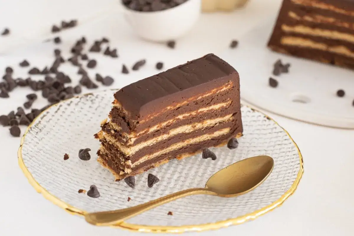 Rebanada de tarta de galletas de la abuela, con capas de galleta y chocolate, decorada con pepitas de chocolate y una cuchara dorada.