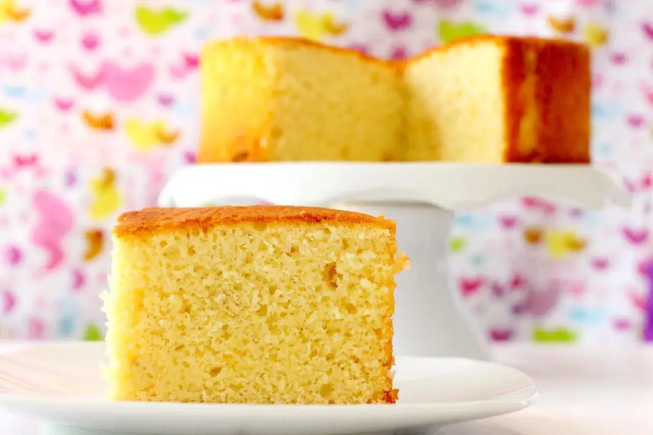 Un trozo de esponjosa coca de yogur sobre un plato blanco, con otra porción en un soporte para pasteles al fondo.