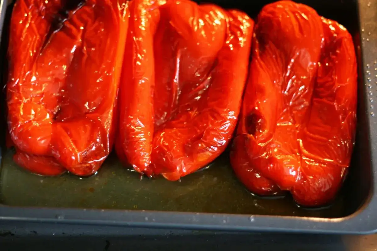 Tres pimientos rojos asados, jugosos y brillantes, reposan en una bandeja con su jugo.