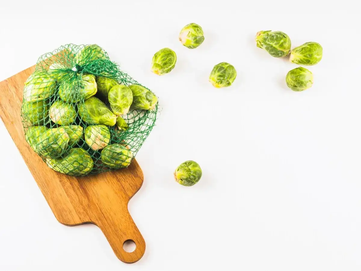Una bolsa de malla verde llena de coles de Bruselas frescas sobre una tabla de cortar de madera. Algunas coles de Bruselas están esparcidas alrededor.