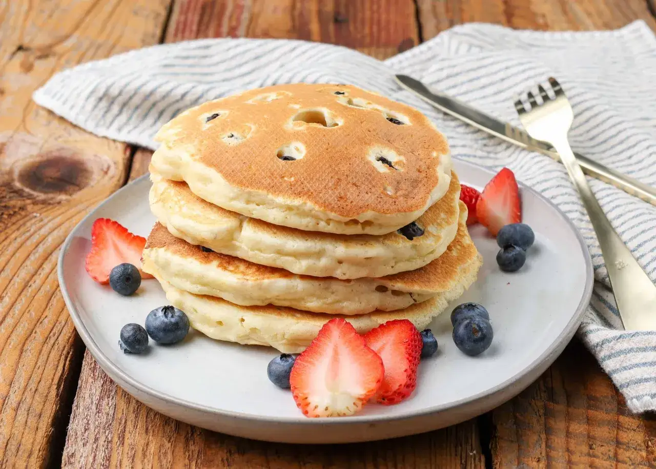 Pila de tortitas americanas con arándanos, fresas y un tenedor dorado.