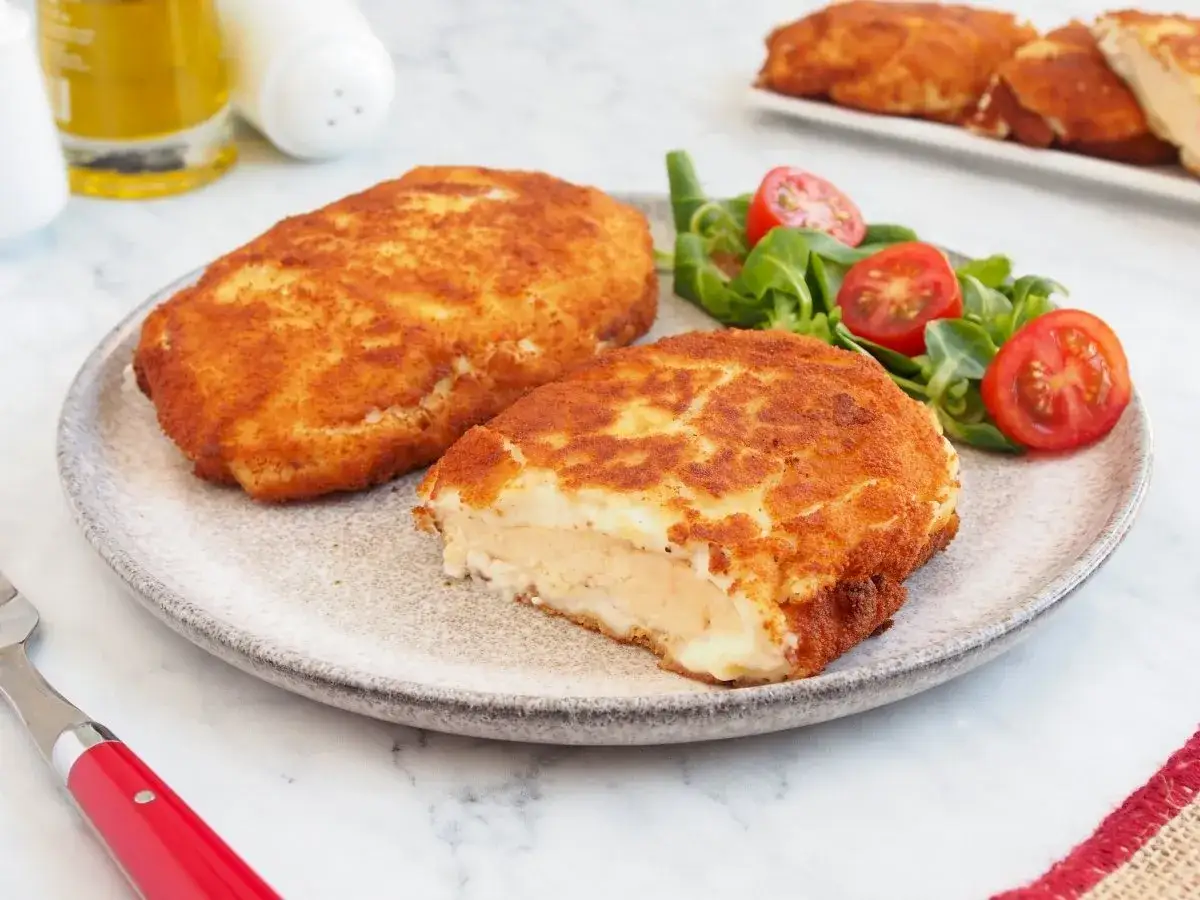 Pechuga villaroy dorada y crujiente, rellena de queso fundido, servida con ensalada fresca y tomates cherry.