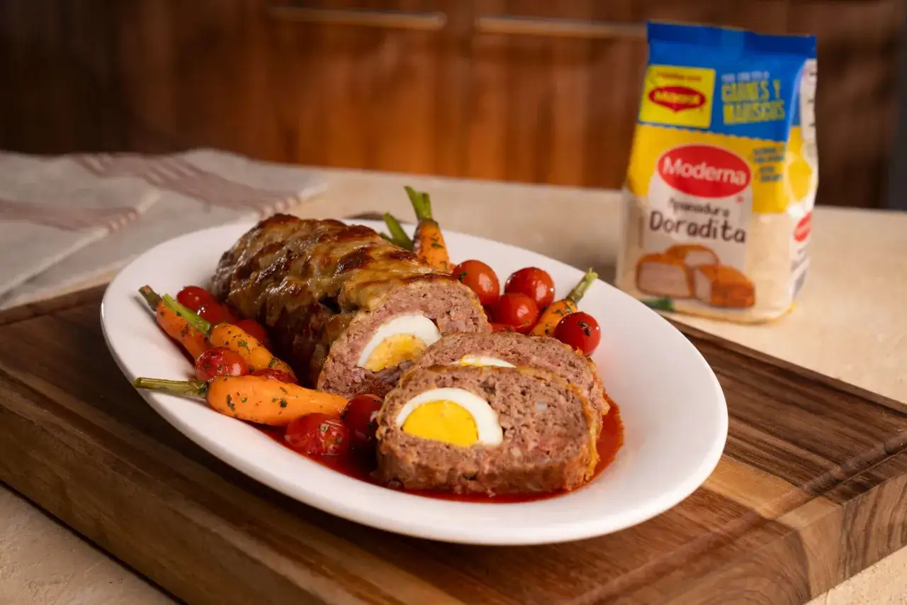 Pastel de carne jugoso con huevo cocido en el centro, acompañado de zanahorias y tomates.