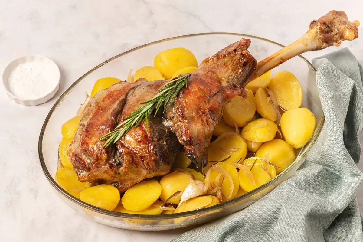 Pierna de cordero al horno dorada y jugosa, servida con patatas asadas y romero en una fuente de cristal.