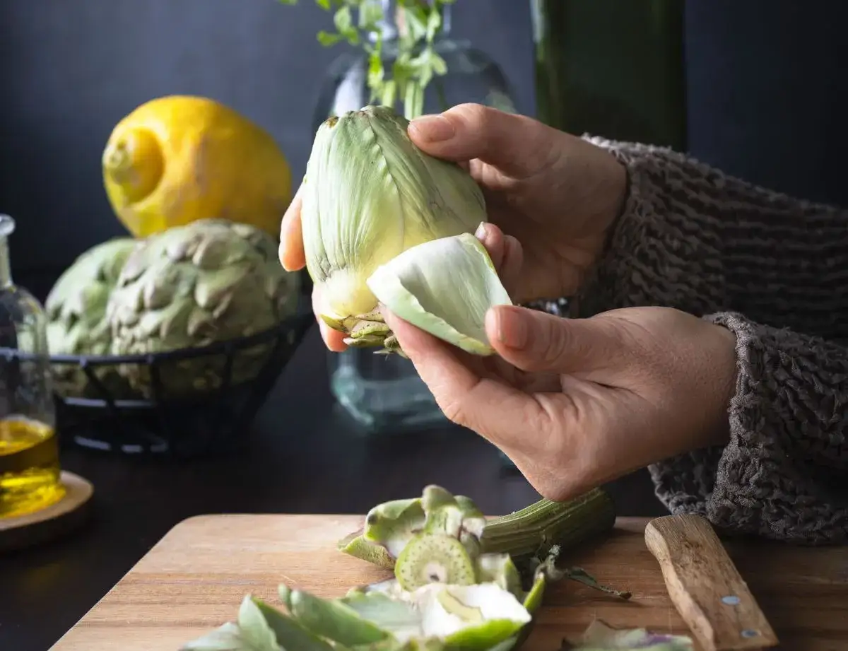 Manos preparan alcachofas para deliciosas recetas. Limón y aceite de oliva completan la escena.