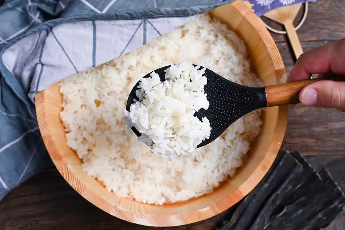Arroz para sushi listo para la receta. Una cuchara negra con mango de madera saca arroz blanco de un cuenco de madera.