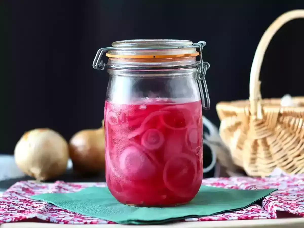 Frasco de vidrio con cebolla encurtida, rodajas rosadas vibrantes sumergidas en líquido. Dos cebollas crudas al lado.