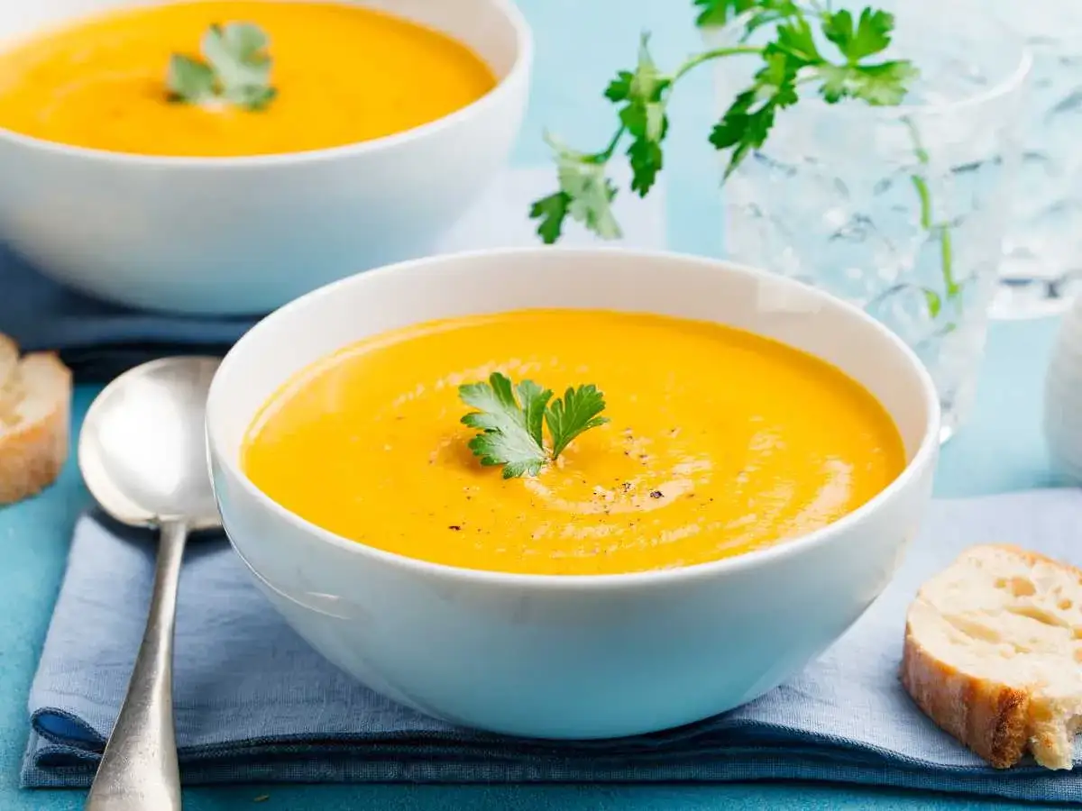 Dos tazones de cremoso pure de calabaza, decorados con perejil, acompañados de pan crujiente y agua con hielo.