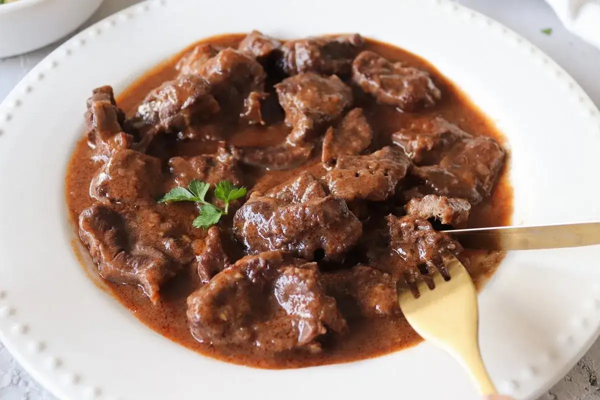 Tierno jarrete de ternera estofado en una rica salsa, servido en un plato blanco con perejil. Un tenedor dorado corta un trozo jugoso.